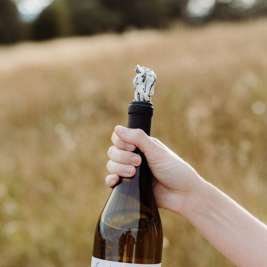 A person holds a bottle of wine in a real outdoor field setting. The bottle has a dark brown glass body, a cream-colored label with black text and illustrations, and a black capsule with a white stopper made of wax