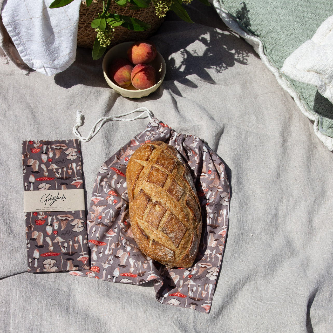 A patterned reusable bread bag with a drawstring closure, shown holding a loaf of bread on a light gray linen blanket. A matching branded bag with a paper band reading 'Breadlovers' is placed beside it, with peaches and greenery in the background.