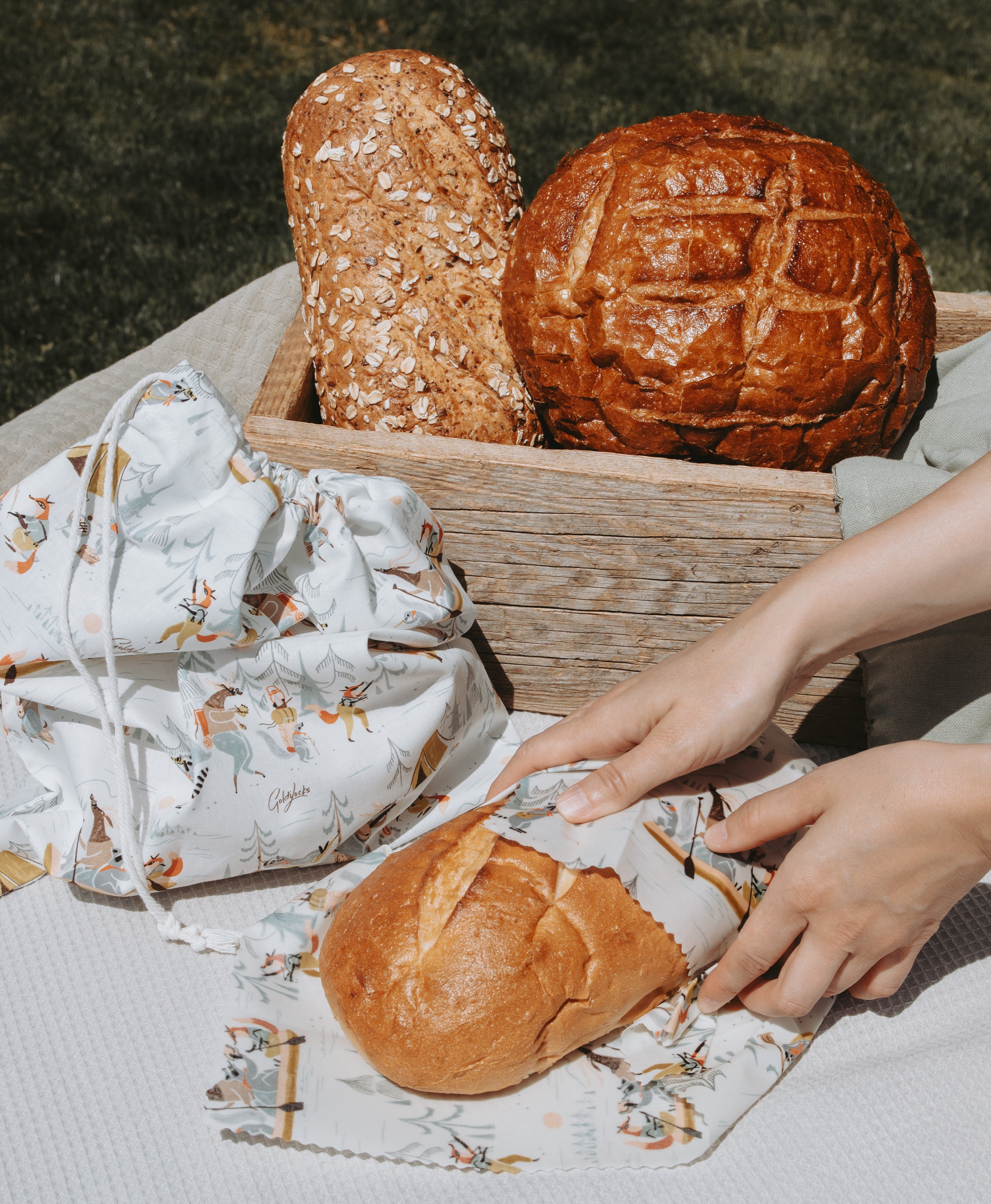 A lifestyle shot of a rustic bread basket containing two loaves of bread, with a person's hands unwrapping a matching animal-patterned beeswax wrap from a loaf in the foreground. A matching drawstring bag is also visible on the left, all arranged on a white textured blanket outdoors.