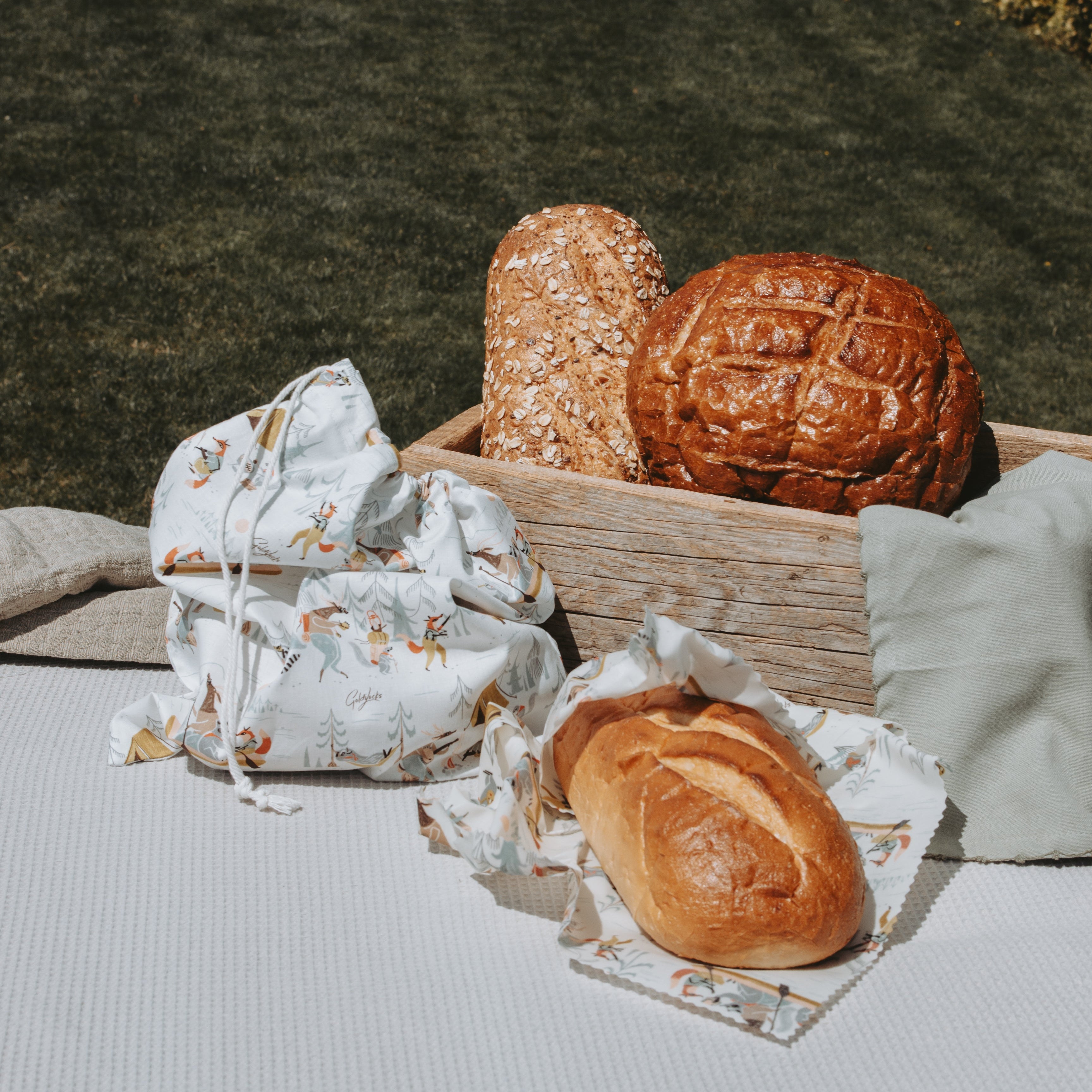 A styled outdoor picnic scene on a white tablecloth with a wooden crate holding two loaves of bread, a white drawstring bag with a small outdoors print and a reusable beeswax food wrap for bread in the same print, and a folded light green cloth, with a grassy lawn in the background.
