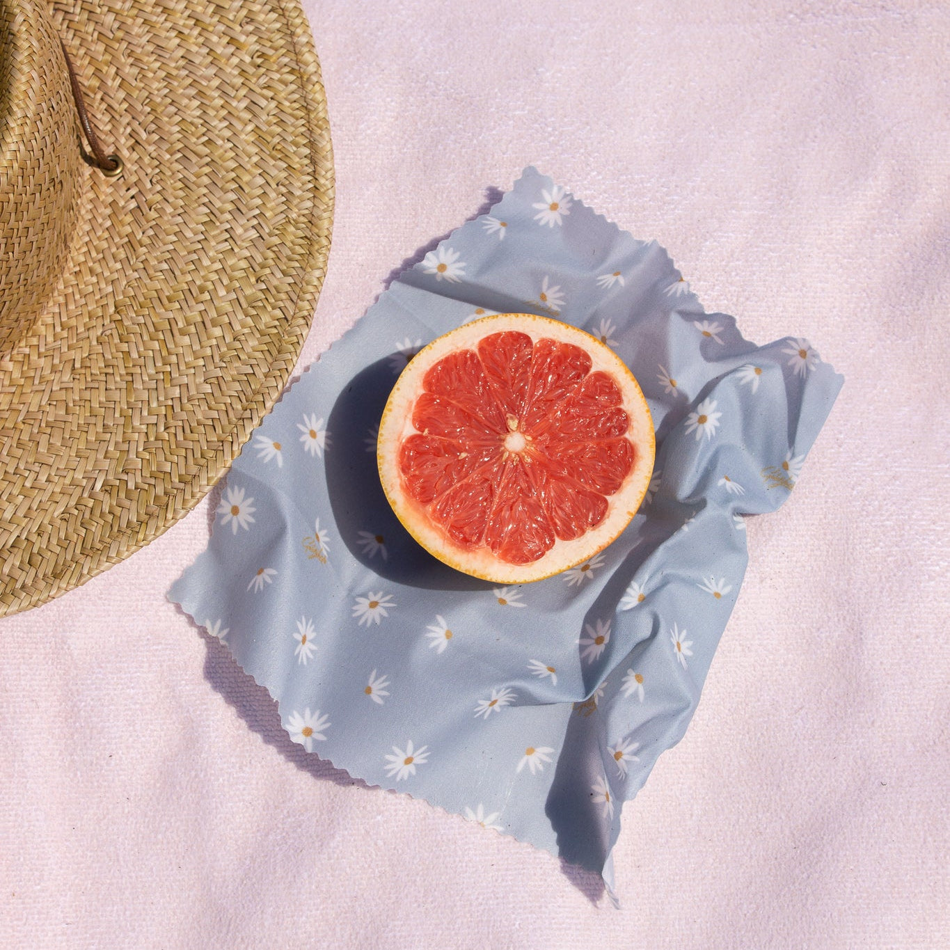 Half of a grapefruit on a blue floral fabric with a straw hat beside it on a light pink background