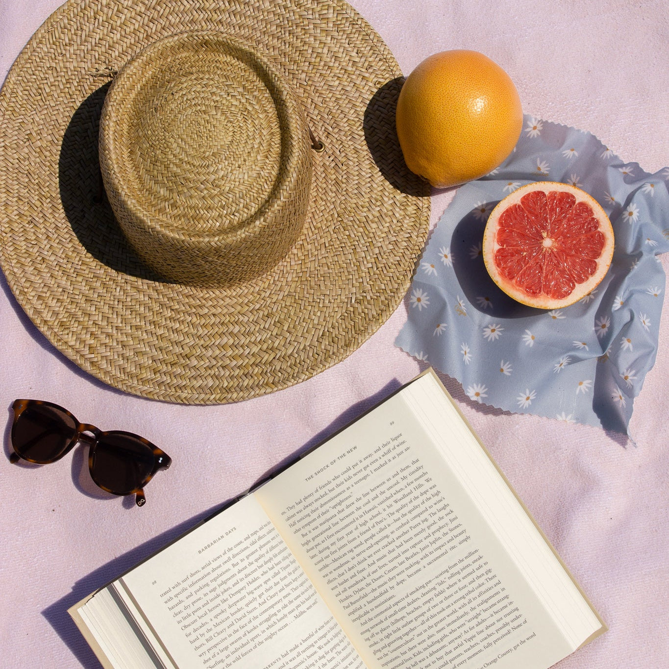 Sun hat, sunglasses, open book, orange, and grapefruit on a light purple surface