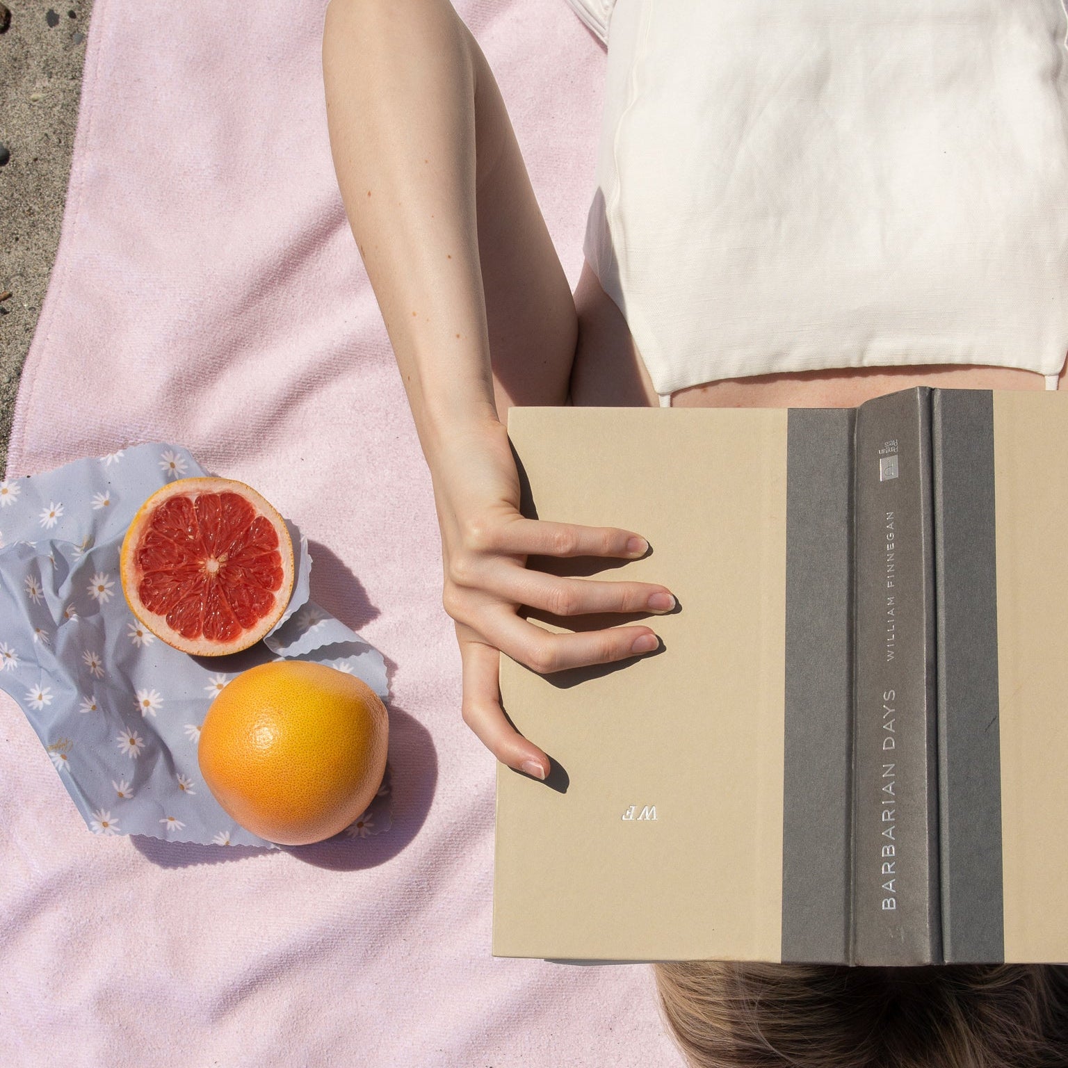Person lying on a pink blanket with a book, grapefruit, beeswax wrap and orange.