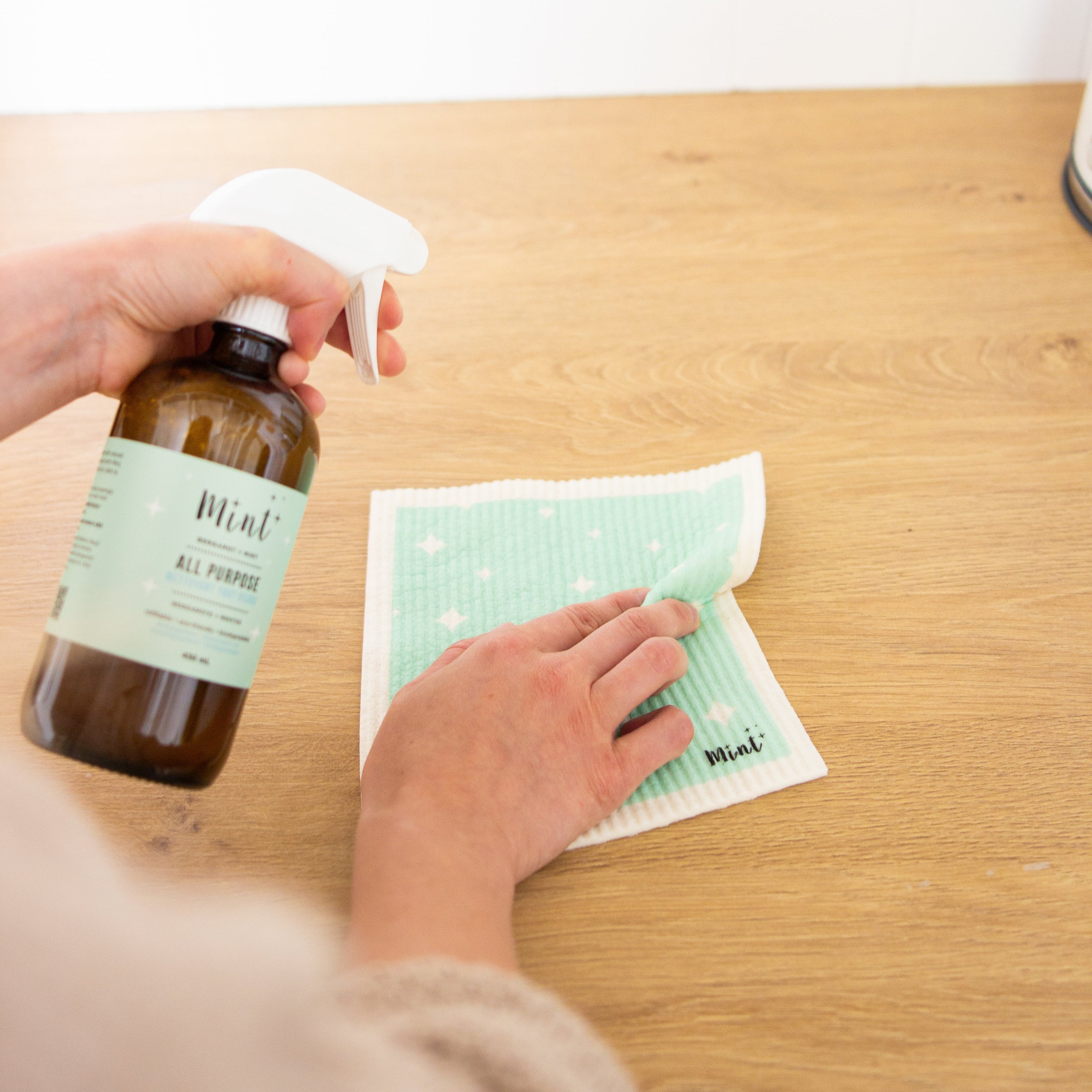 Person cleaning a wooden surface with a mint cleaning spray bottle and dishcloth
