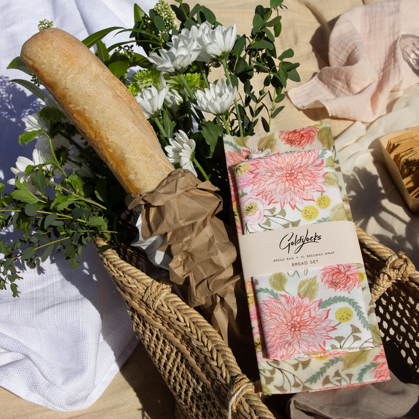 A woven wicker basket filled with a long baguette, white flowers, greenery, and a rectangular box wrapped in pink floral paper with the brand name 'Ghigliote' visible, set on a light fabric surface with draped white and blush textiles.
