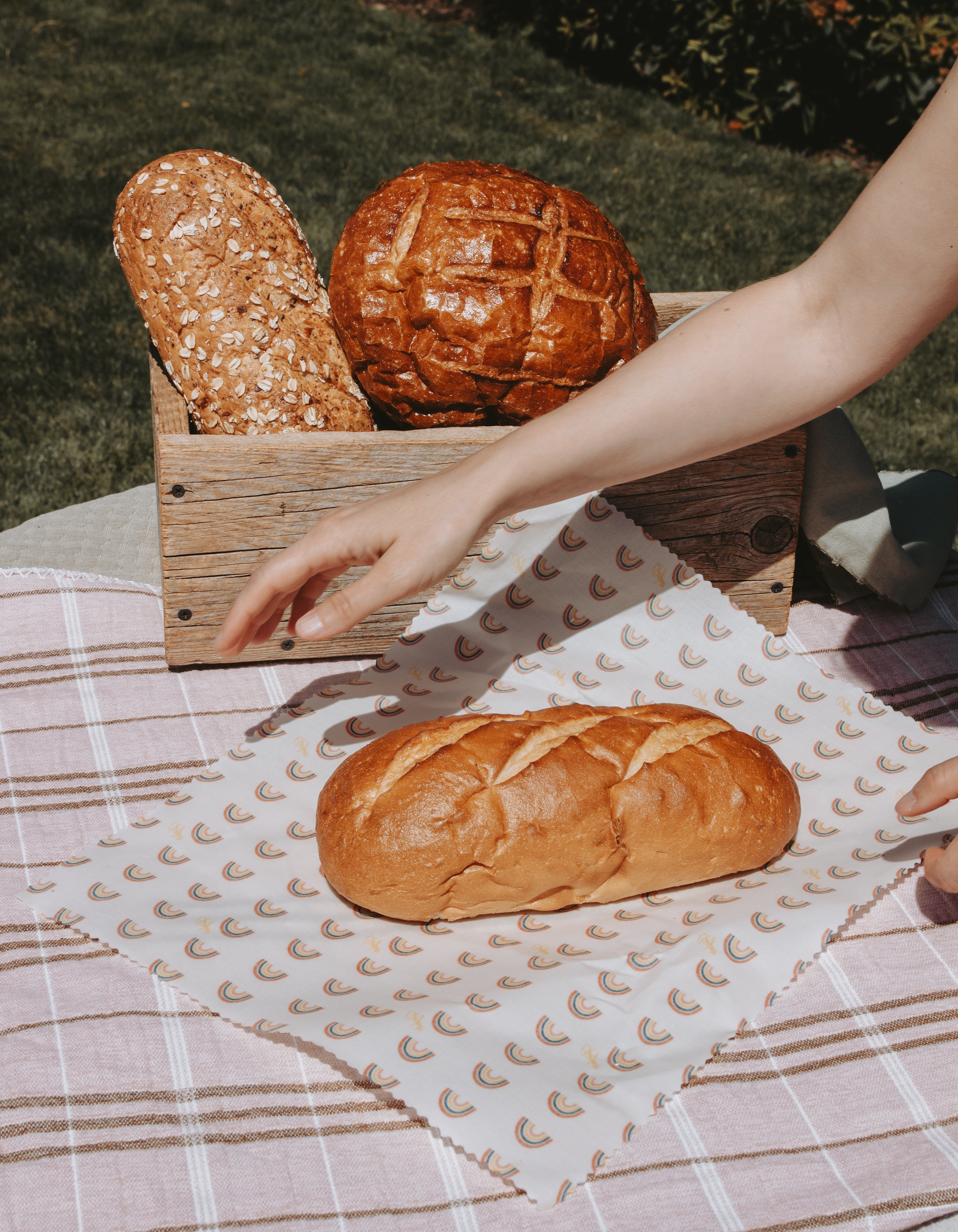 Freshly baked artisanal bread loaves displayed outdoors on a picnic blanket, with one loaf in a wooden crate and another on a patterned beeswax wrap in the foreground. A person's arm is reaching toward the bread.