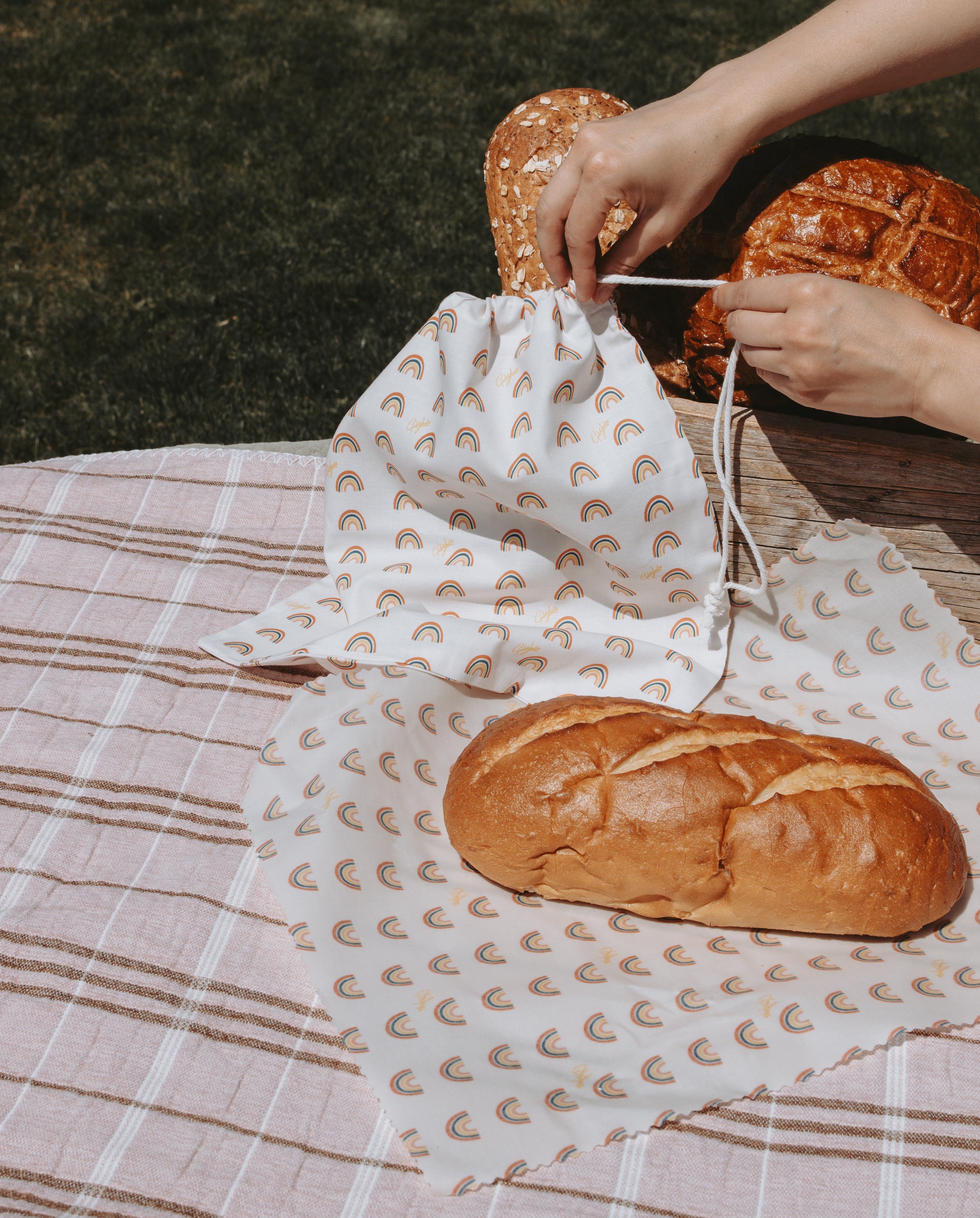 A reusable fabric bread bag and a reusable beeswax food wrap with a rainbow pattern is being filled with two loaves of bread by hands, set on a pink and white plaid picnic blanket outdoors.