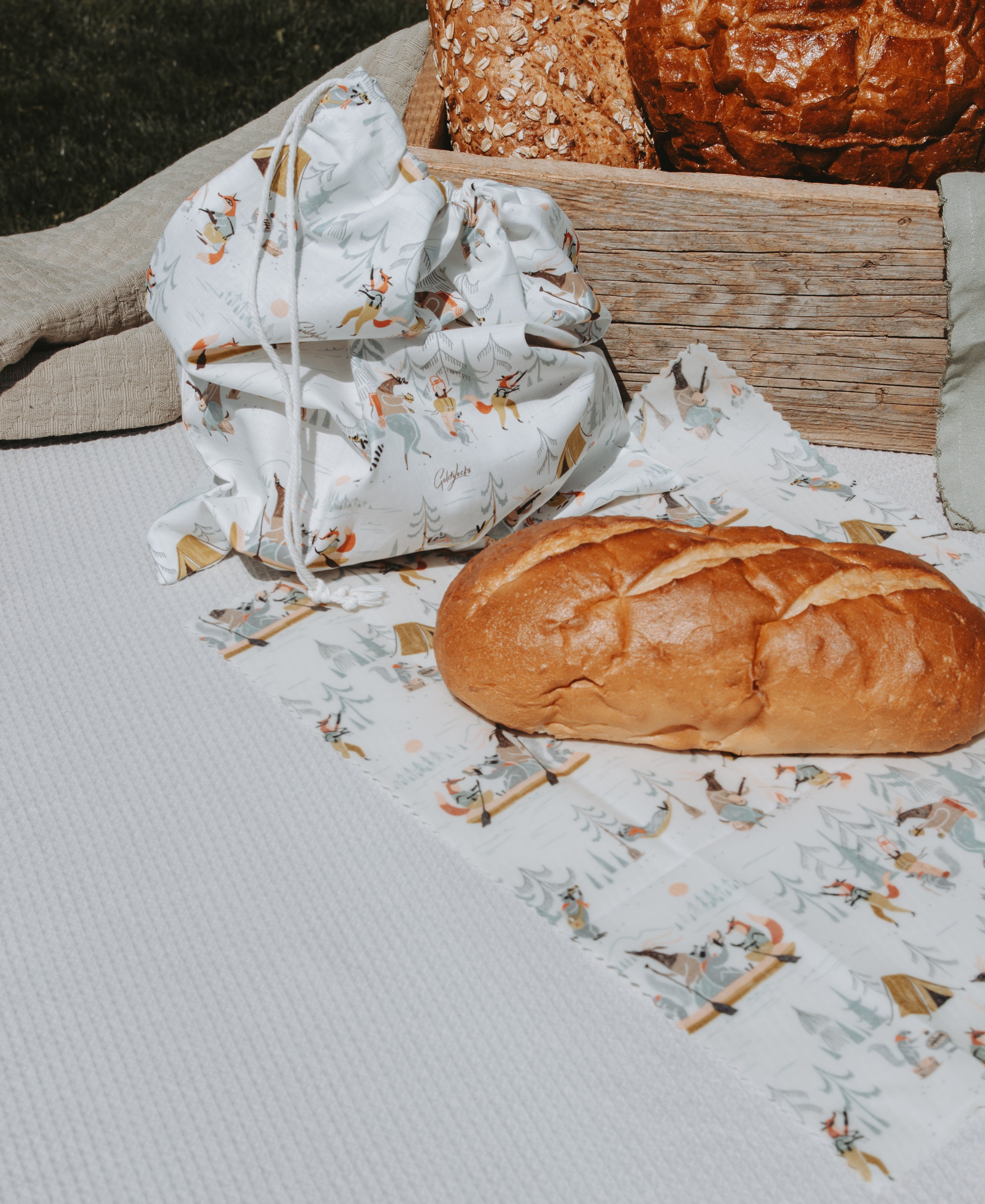 A white textured picnic blanket with a reusable beeswax food wrap with a printed pattern of small animals and trees, styled with a loaf of bread and a matching drawstring bag.
