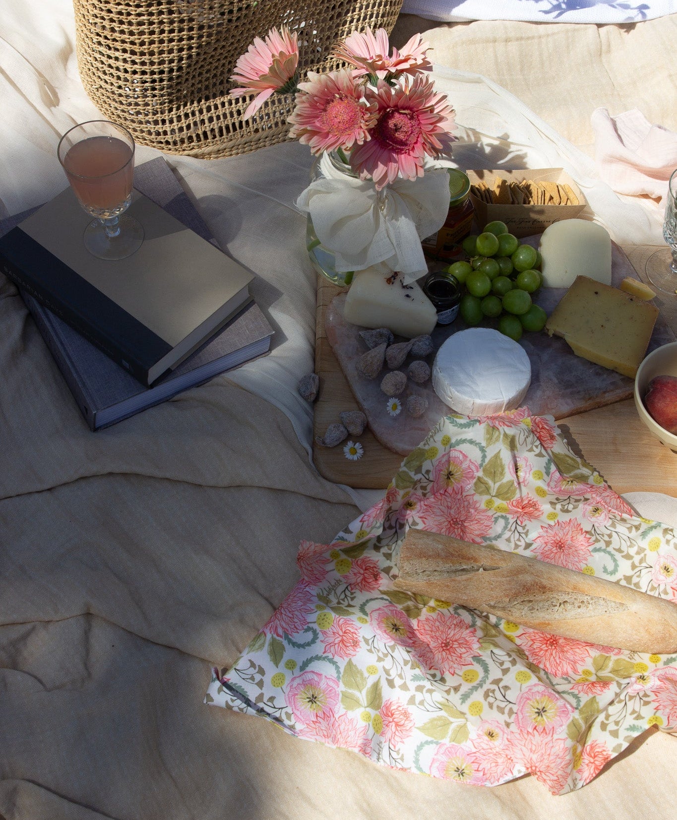 A styled picnic scene on a beige blanket with a woven basket, flowers, grapes, cheese, books, and a floral napkin in warm natural light. French baguette on top of a reusable beeswax food wrap with pink floral pattern