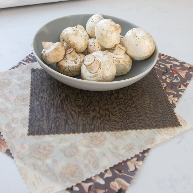 a bowl of white mushrooms placed on top of 3 different beeswax wraps: a small wood grain wrap, a medium floral print wrap and a large mushroom print wrap